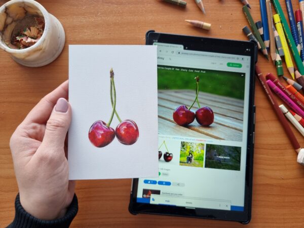 A hand holding a cherry illustration next to a tablet with the reference drawing.