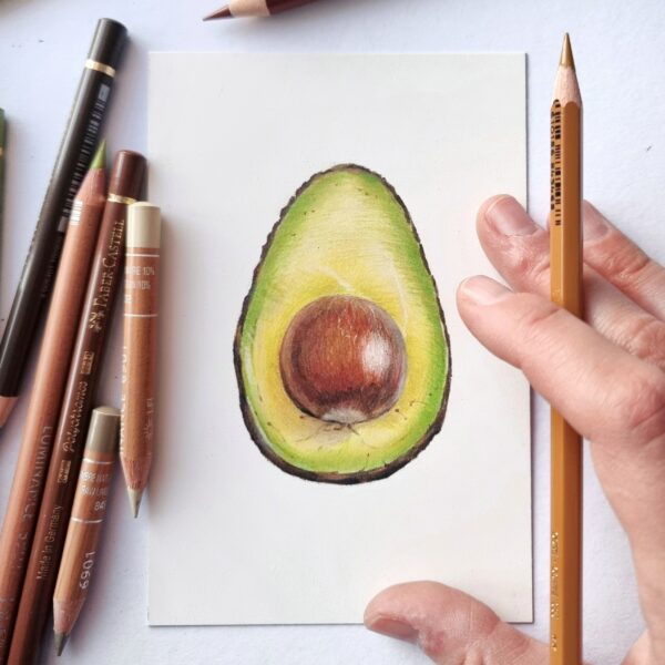 Avocado drawing on a table, coloured pencils on one side, hand holding a pencil on the other side.